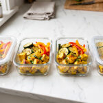Quattro contenitori in vetro per il meal prep allineati su un piano cucina in marmo, contenenti cubetti di pollo al curry, zucchine grigliate e peperoni rossi e gialli saltati.